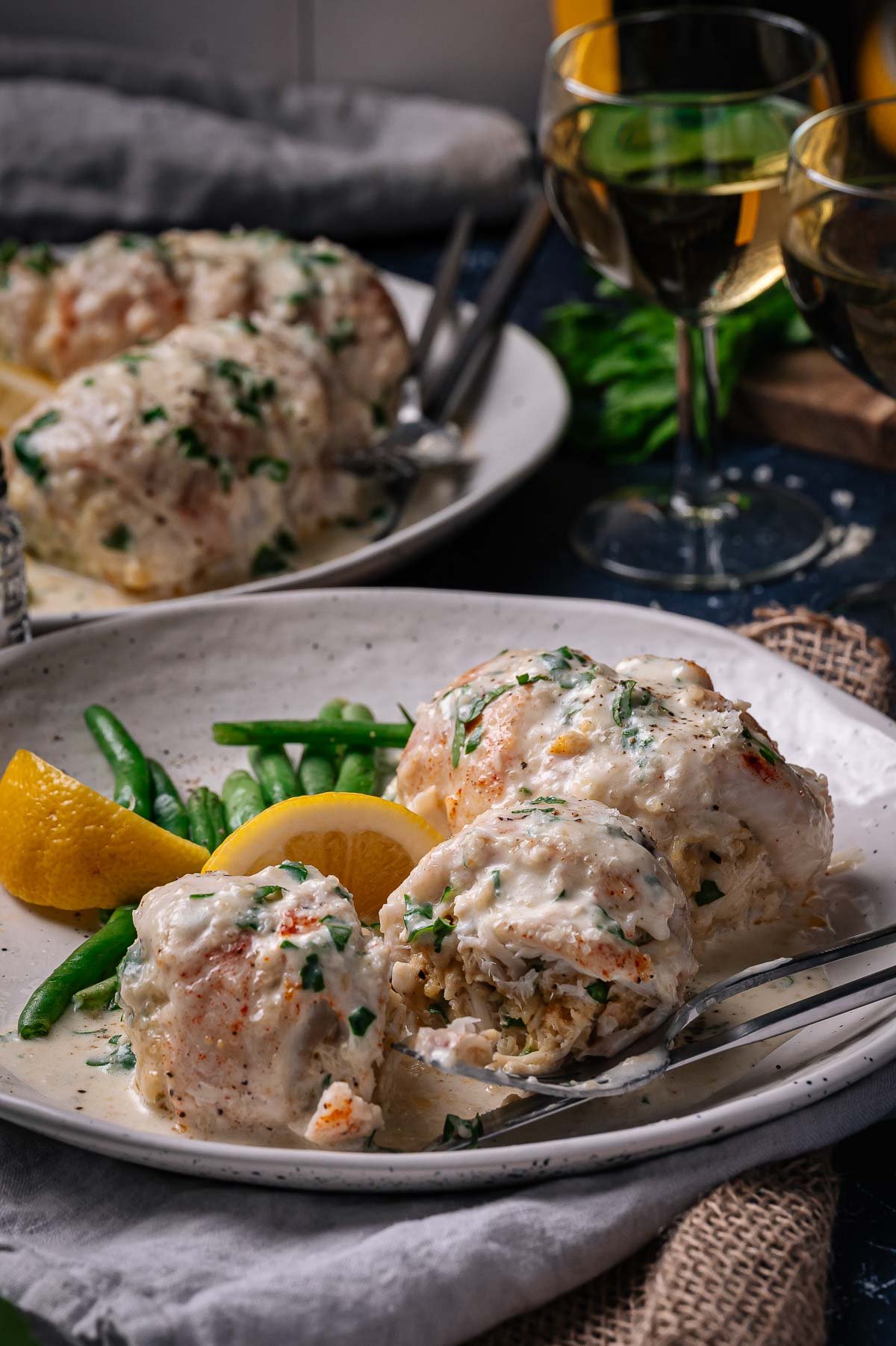 A plate of stuffed flounder topped with creamy white wine sauce, served with green beans and lemon wedges, and two glasses of white wine in the background.