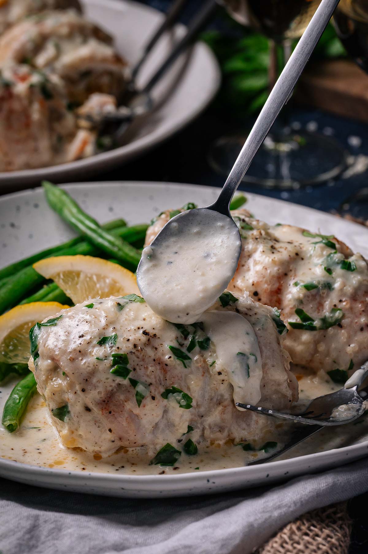 A spoon pours creamy white wine sauce over baked chicken breasts garnished with herbs, served with green beans and lemon slices on a white plate.