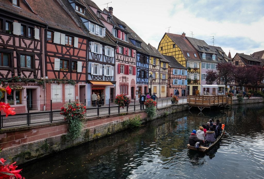 A canal in the city of Colmar, France, with multicolored half-timbered homes along the water. A small boat with people in it floats in the water.