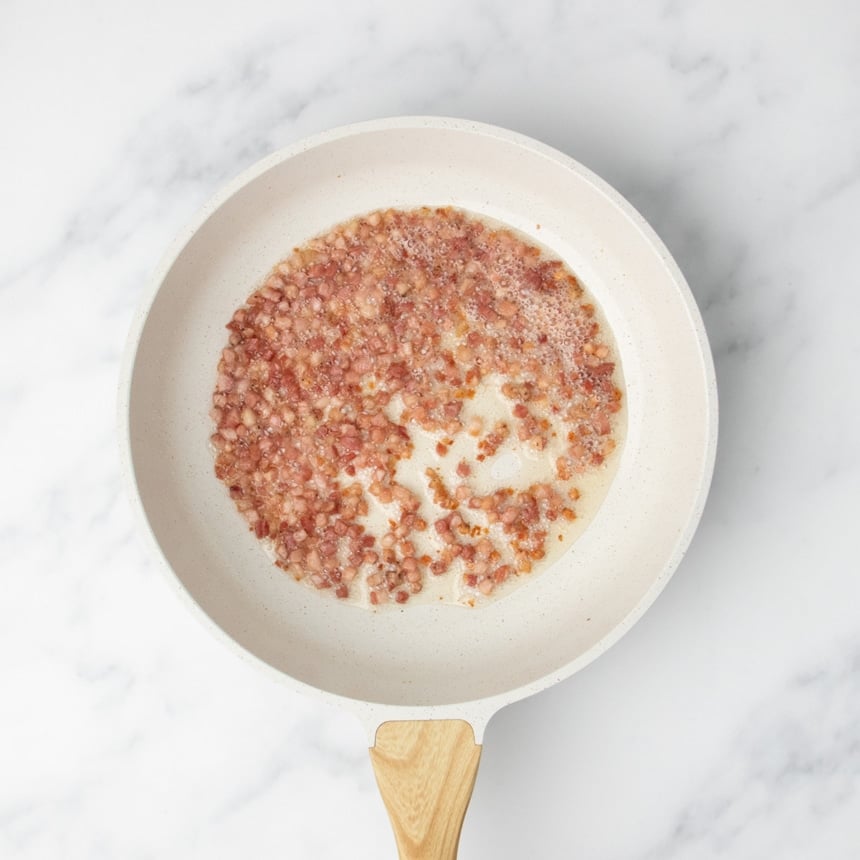 Diced pancetta cooking in a white skillet on a marble surface.