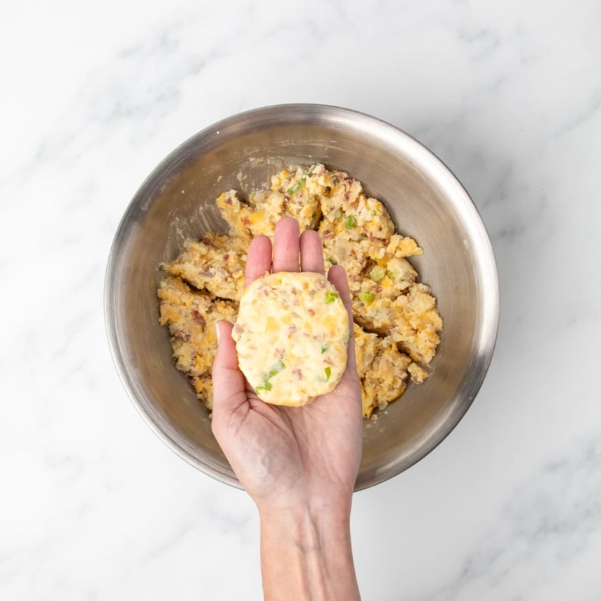 A hand holding the ball of mashed potato mixture flattened into a patty above a bowl of the same mixture.