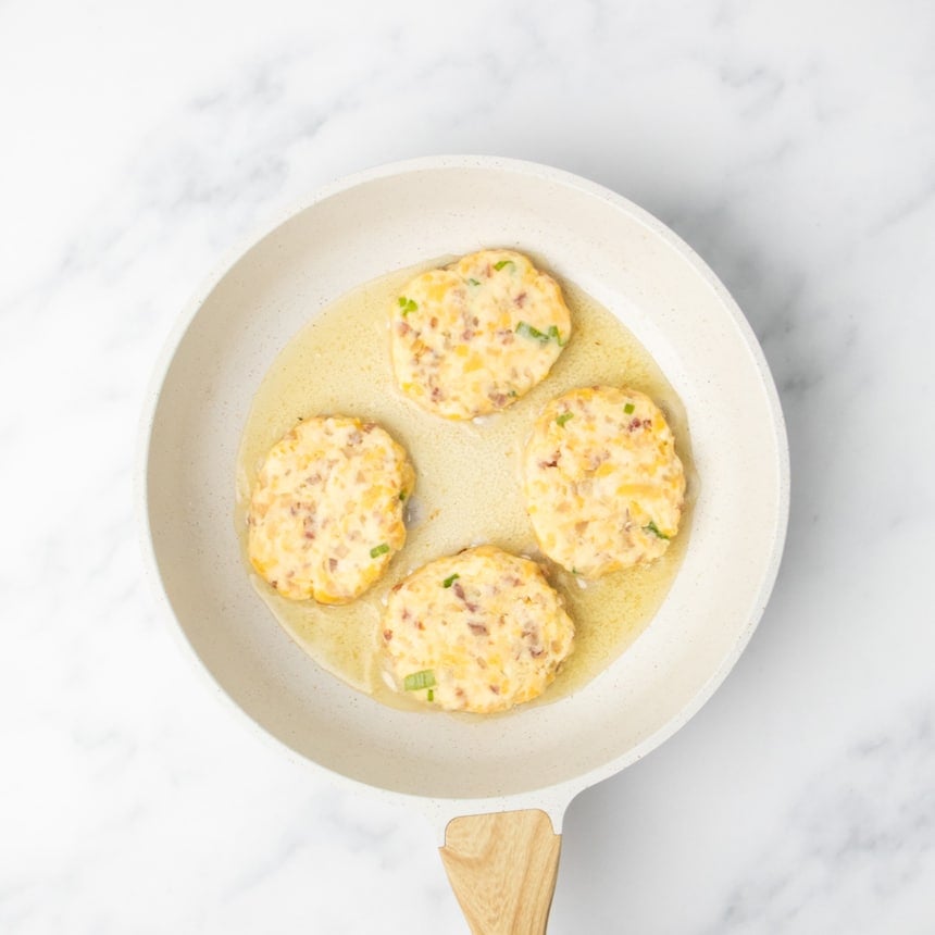 Four uncooked patties of mashed potato mixture frying in oil in a white skillet on a marble surface.
