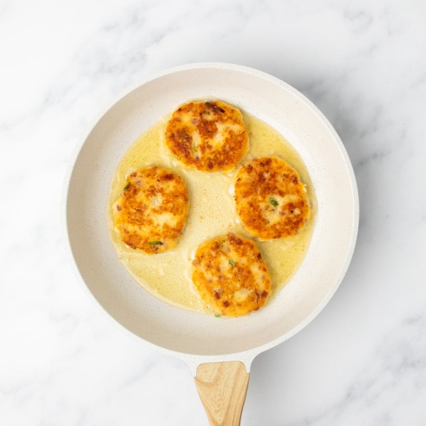 Four golden brown fried potato patties cooking in oil in a white skillet on a marble surface.