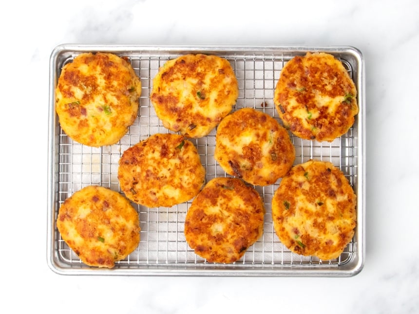 Eight browned potato patties cooling on a wire rack set over a small baking sheet, which is lined with white paper towels.