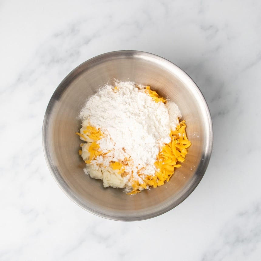 Grated yellow cheddar cheese, flour, and mashed potatoes in a stainless steel mixing bowl on a marble surface.