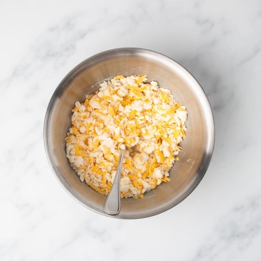 Flour, mashed potatoes and grated cheddar mixture roughly combined in a stainless steel bowl with a mixing spoon.