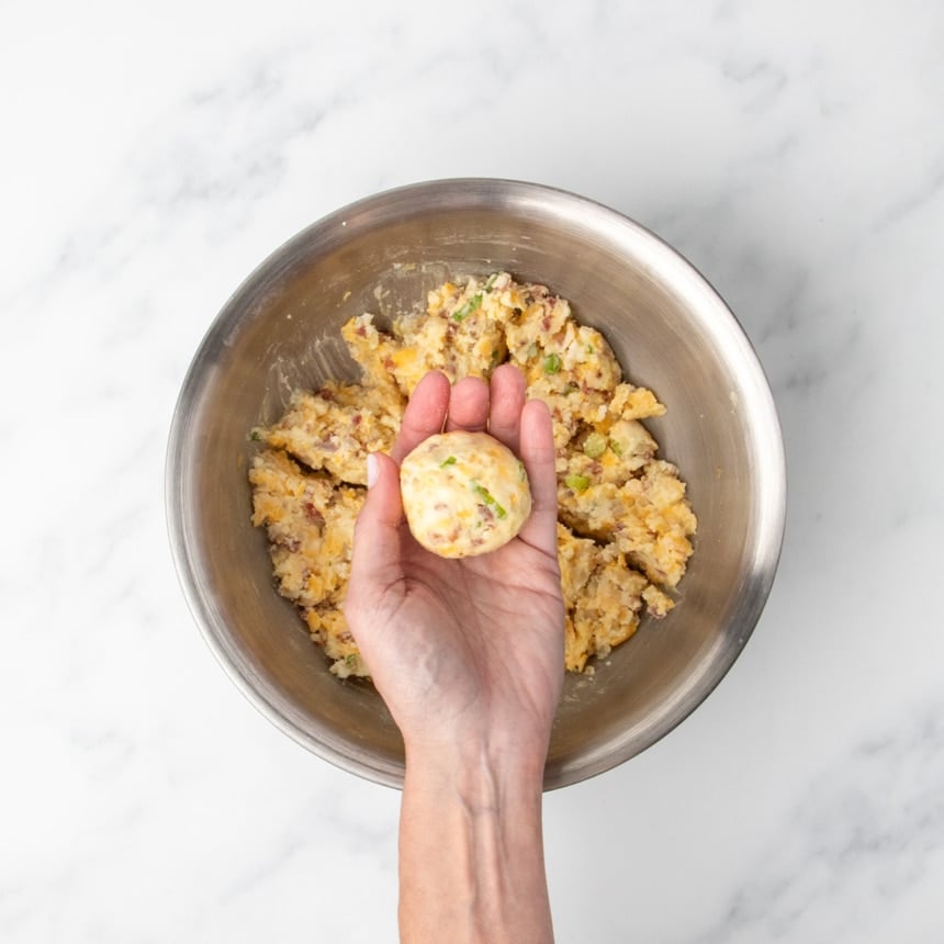 A hand holding a round ball of mashed potato mixture above a bowl of the same mixture.