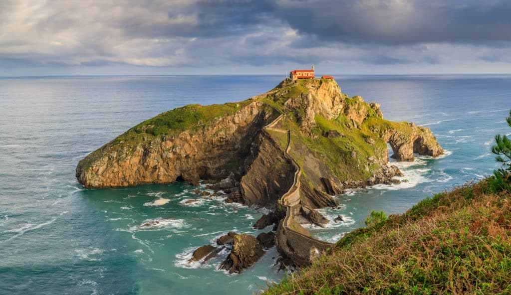 A gorgeous rocky island just off the Basque coast with a little road leading to a building on top.