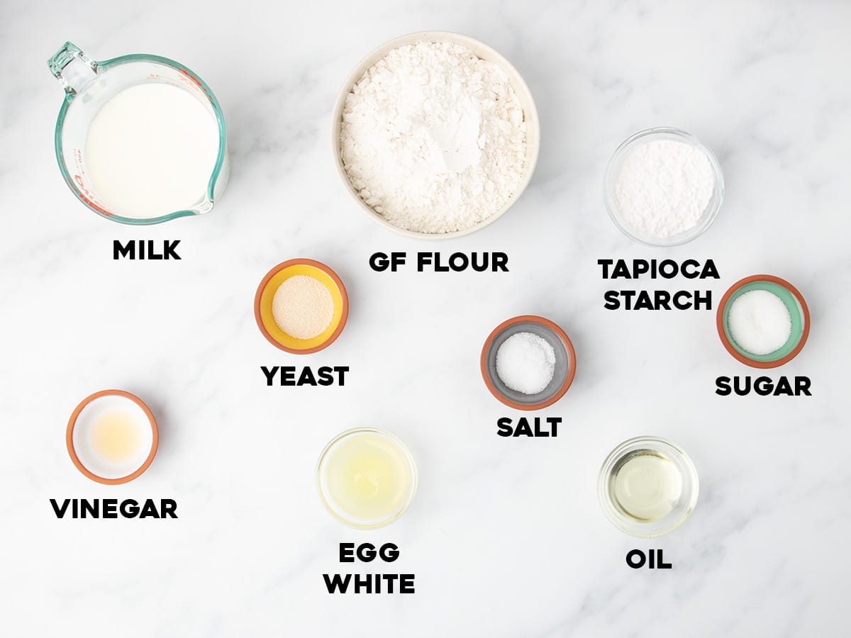 Ingredients for gluten free English muffins arranged on a white marble surface in small bowls and cups, each labeled milk, GF flour, tapioca starch, sugar, salt, yeast, vinegar, egg white, and oil.