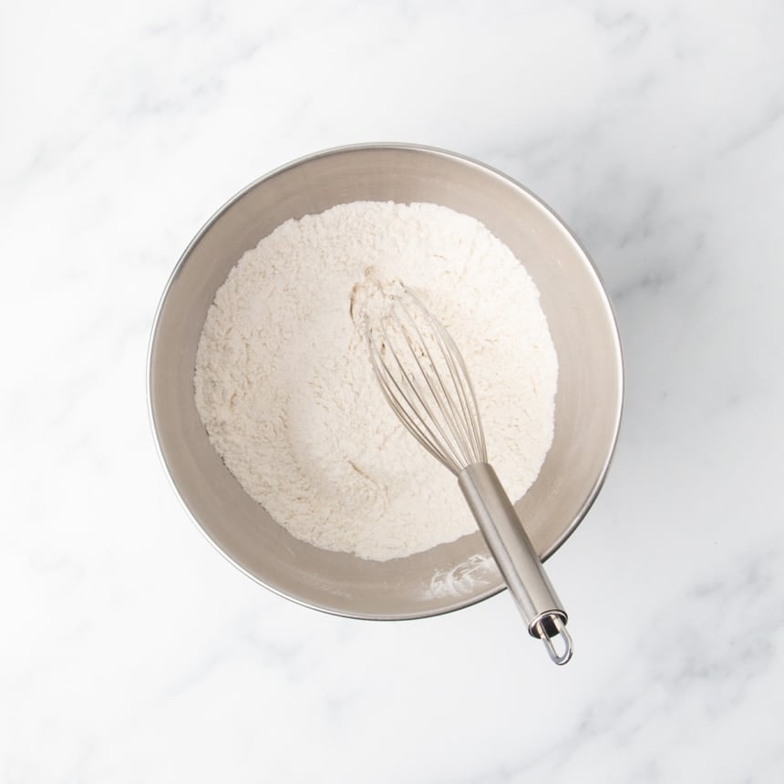 A metal mixing bowl filled with a dry flour mixture with a metal whisk resting inside on a white marble surface.