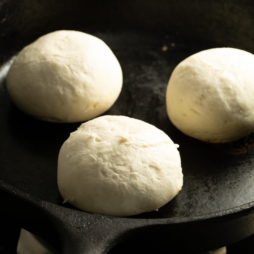 Three dough rounds cooking directly in a cast iron skillet, round open rings removed, with smooth tops and light color.