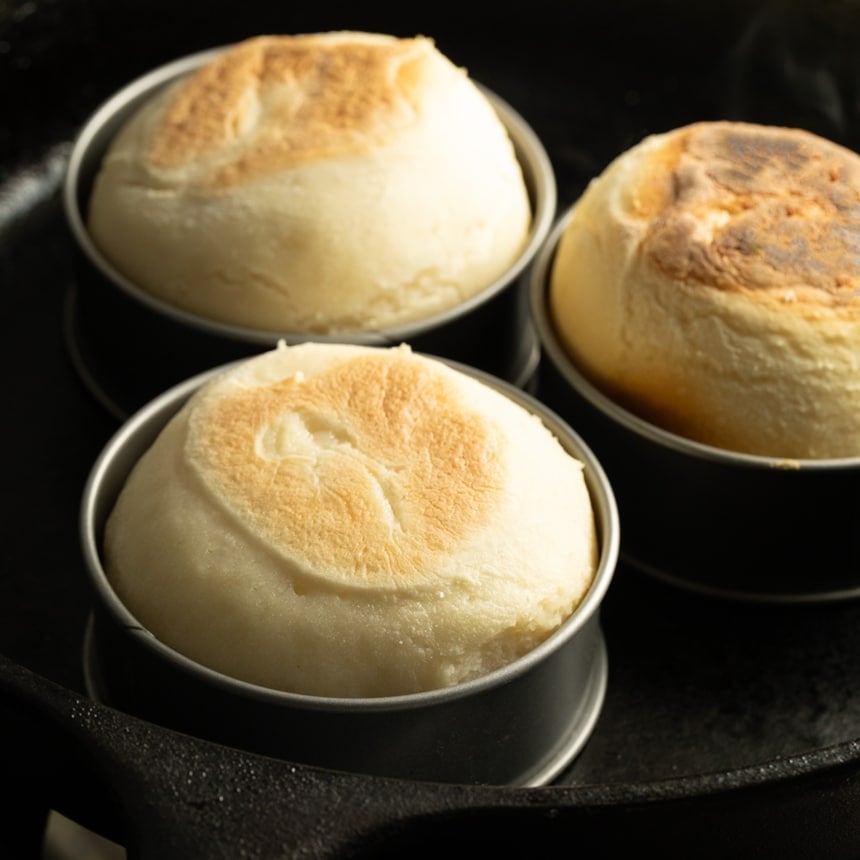 Three fully-risen English muffins in metal molds in a cast iron skillet, with tops more deeply browned and slightly uneven in color.