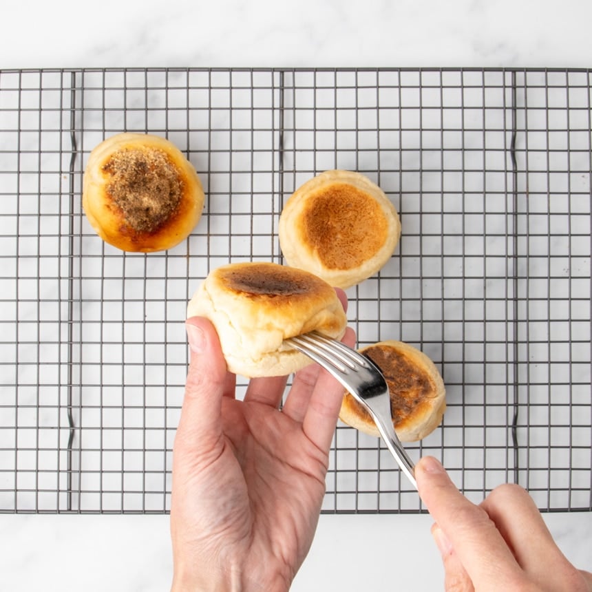A hand using a fork to pierce the side of a browned English muffin while four cooked muffins rest on a wire rack.