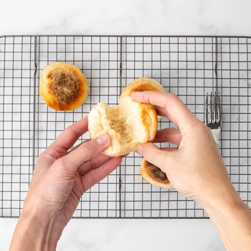 Two hands pulling apart a cooked English muffin to reveal the airy interior, with additional muffins and a fork on a wire rack in the background.