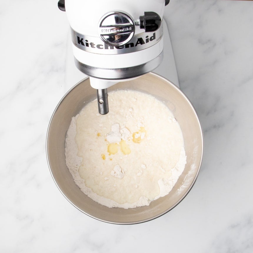 A stand mixer bowl containing flour with milk, vinegar, oil and other wet ingredients just added on top, before mixing.