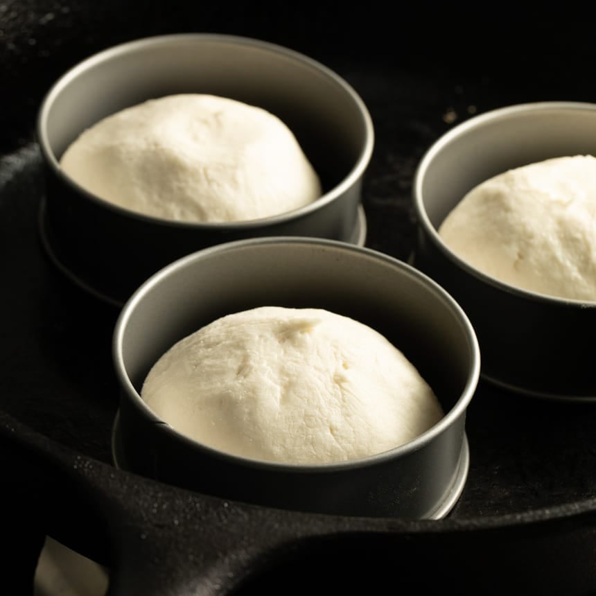 Three portions of unrisen, raw dough in round metal molds placed in a cast iron skillet.