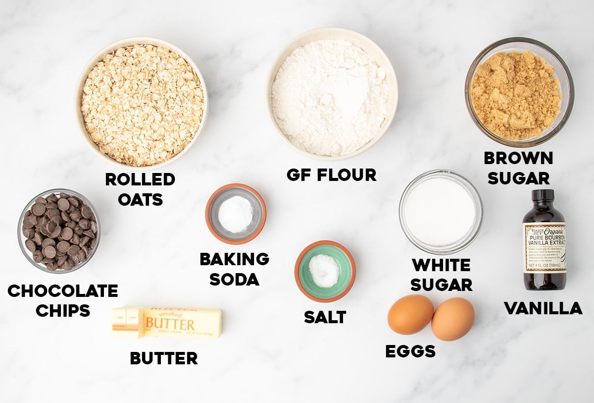Bowls and small dishes on a white surface containing rolled oats, gluten free flour, brown sugar, white sugar, baking soda, salt, butter, eggs, chocolate chips, and a bottle of vanilla extract labeled with text.