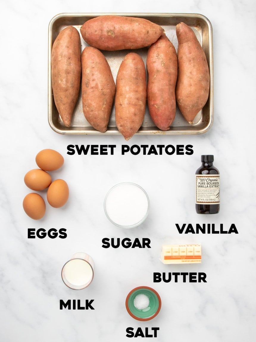 A tray of six whole raw sweet potatoes surrounded by bowls and containers holding eggs, sugar, milk, salt, butter, and vanilla extract, all labeled with bold text on a white marble surface.