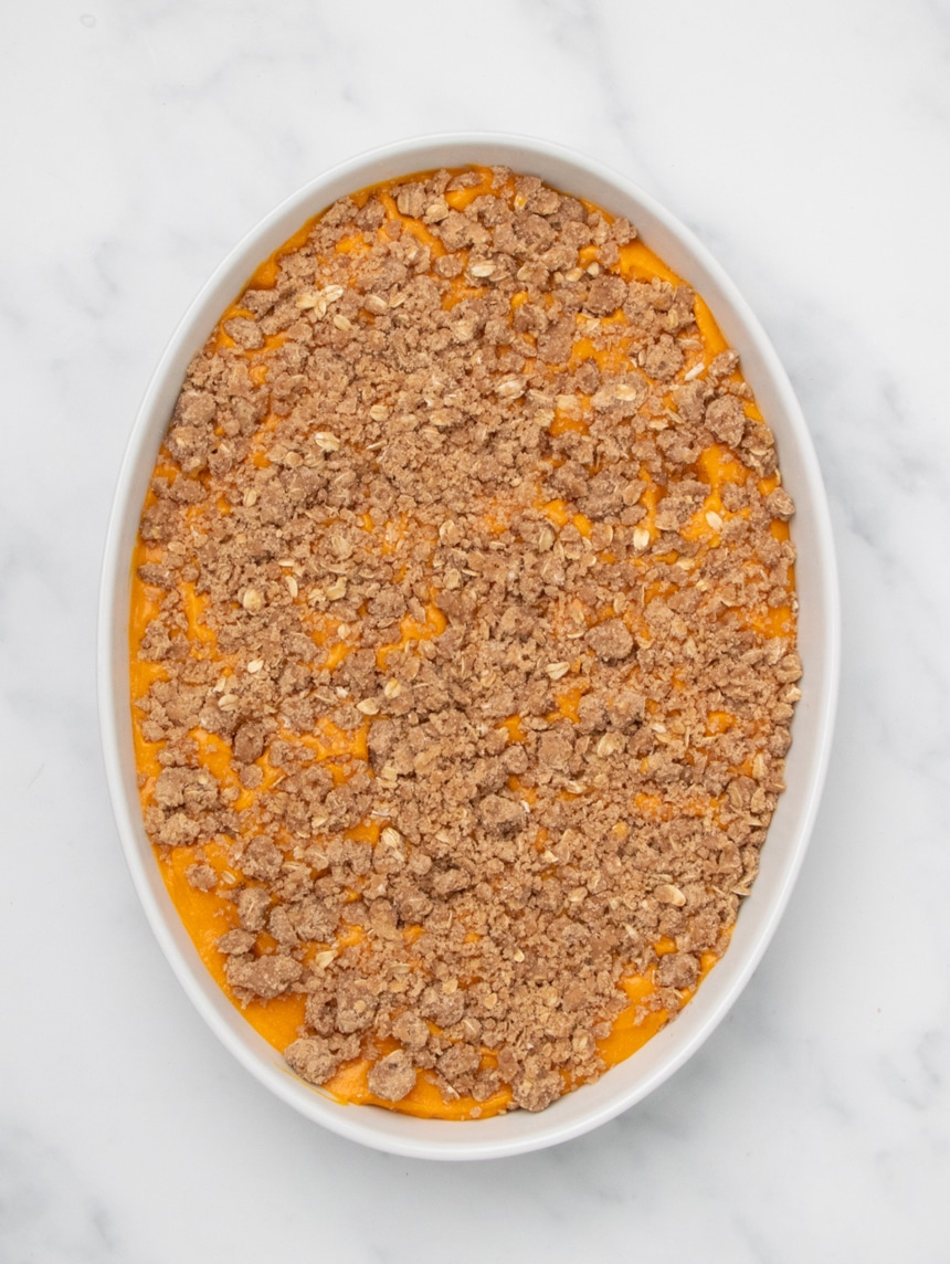 A white oval baking dish filled with orange sweet potato filling topped with an even layer of raw crumb mixture on a white marble surface.
