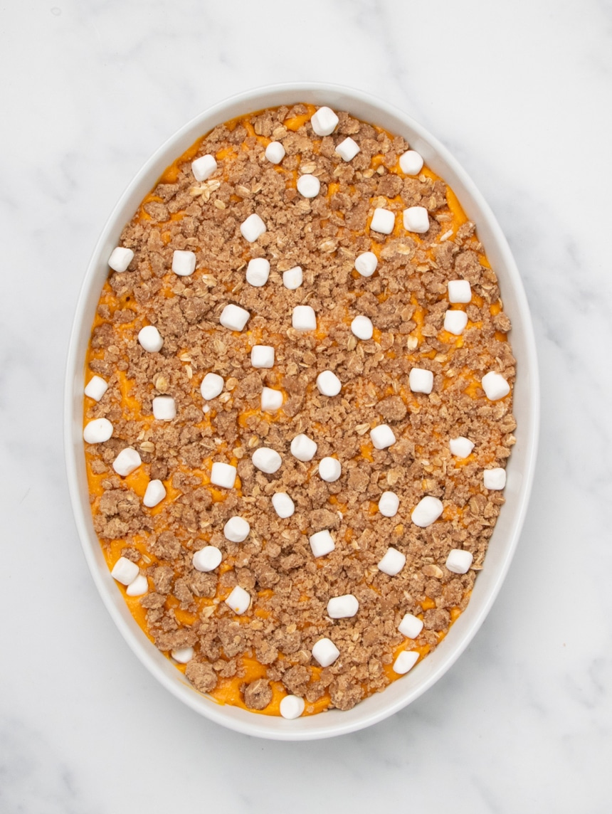 A white oval baking dish filled with orange sweet potato filling topped with an even layer of raw crumb mixture and scattered small white marshmallows, unbaked.
