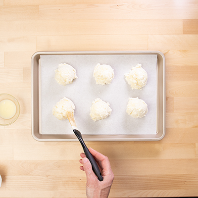 Hand brushing pastry brush with melted butter on raw drop biscuits on paper lined tray.