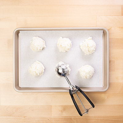 Ice cream scoop with mounds of drop biscuits on white paper on baking tray.