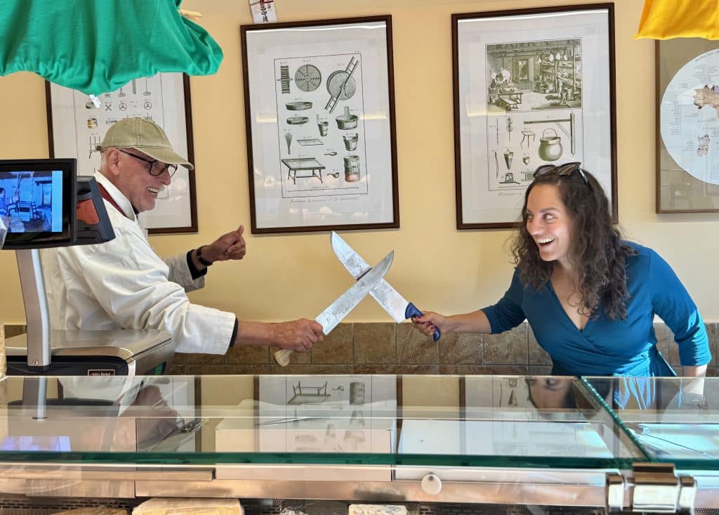 Kate and an older male cheese shop owner standing behind the counter of cheeses, crossing knives like they're sword-fighting.