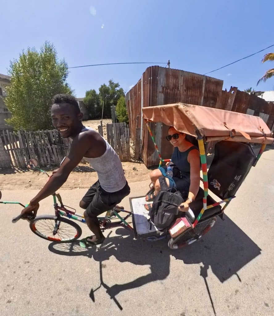 Kate riding in a rickshaw with a roof, called a pousse-pousse, driven by a driver on a bicycle attachment. They both smile big for the camera.