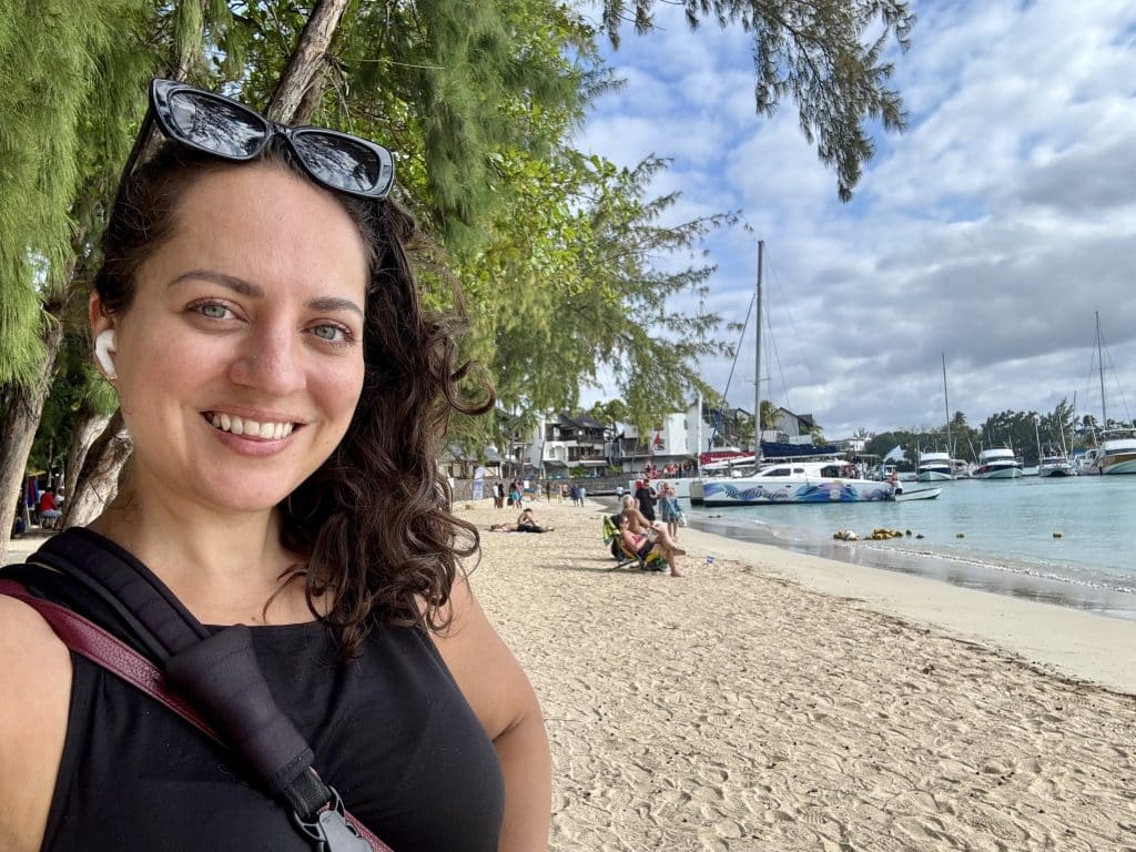 Kate taking a selfie on a beach in Mauritius. Sailboats are in the harbor behind her.