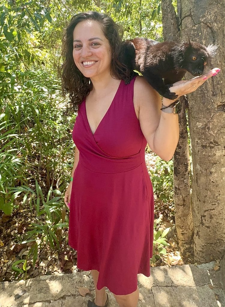 Kate wearing a hot pink dress and a black lemur sitting on her shoulder, eating mashed bananas out of the palm of her hand. She smiles enormously.