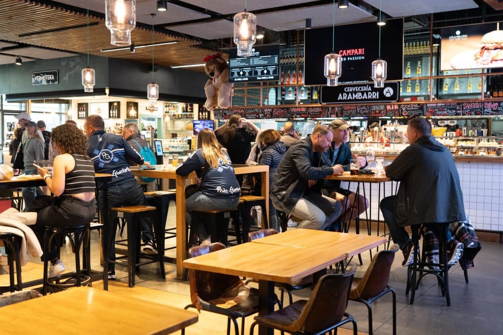 People sitting at high-top tables while others order food from counters in a market.