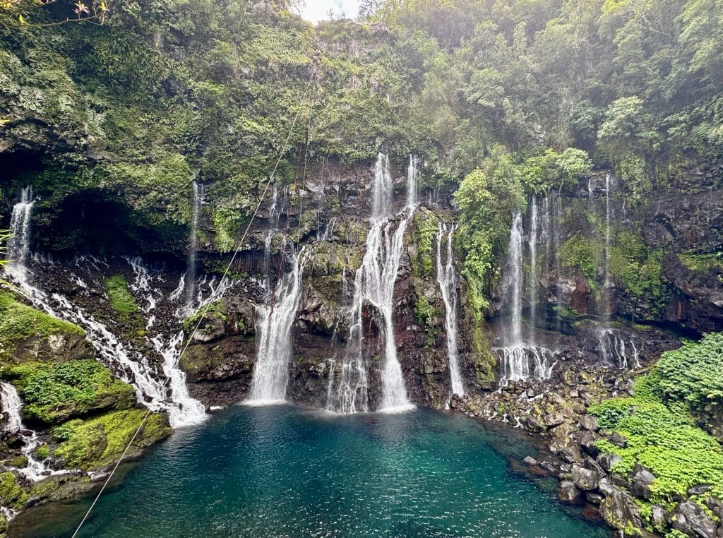 A multi-tiered, bridal veil-style waterfall, with tall green cliffs leading into a deep, clear, teal pool.