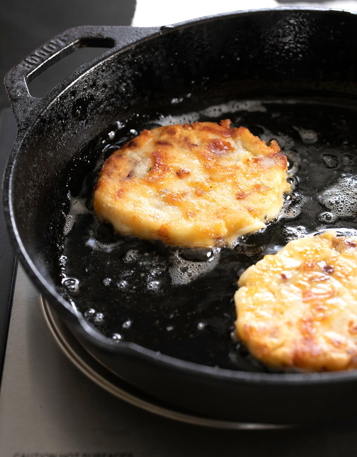 yellow round fried mashed potatoes with brown bits frying in oil in black cast iron skillet