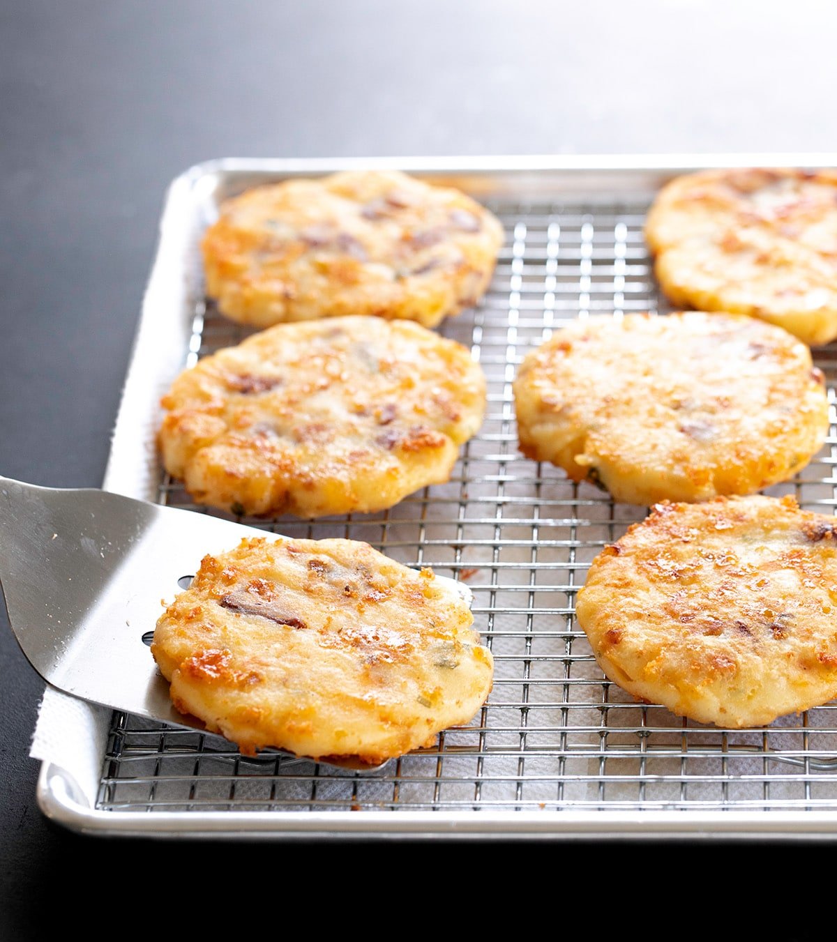 Metal spatula placing fried mashed potatoes on wire rack on top of lined baking sheet