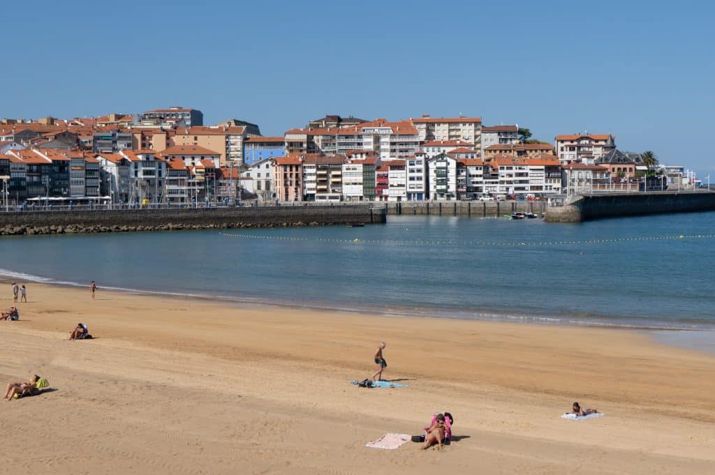 A long sandy beach in the foreground, while in the background you see a small village with lots of square window-filled buildings overlooking a marina and the sea.