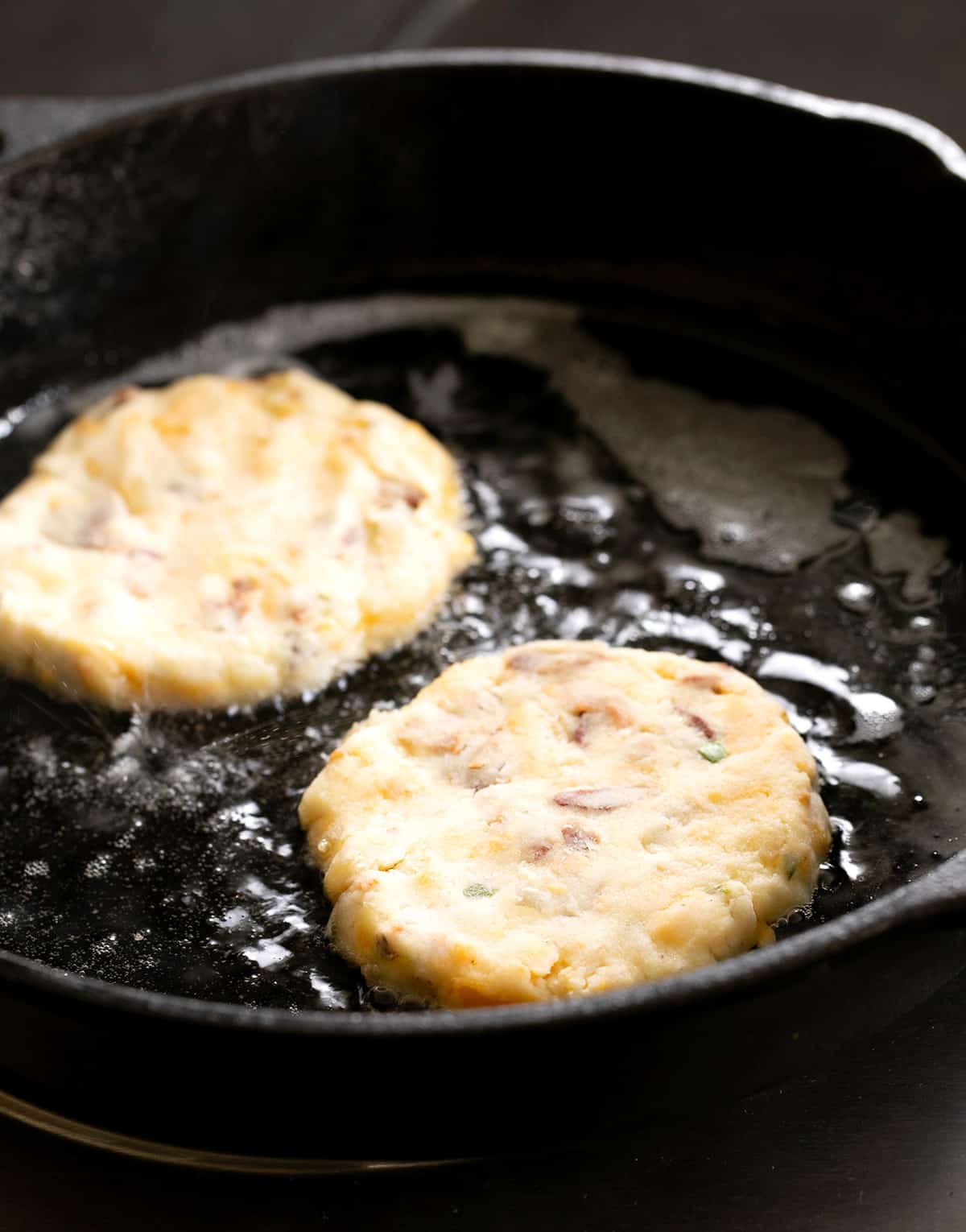 Light yellow unbaked mashed potato cakes beginning to fry in oil in black skillet