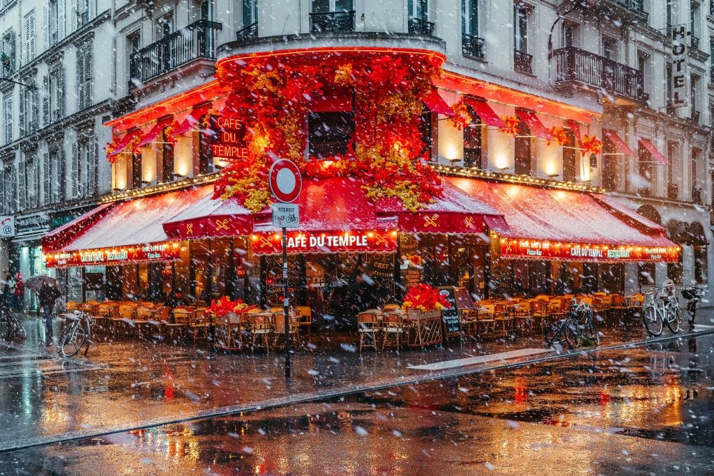 A bright red cafe on a street corner in Paris underneath a snow flurry.