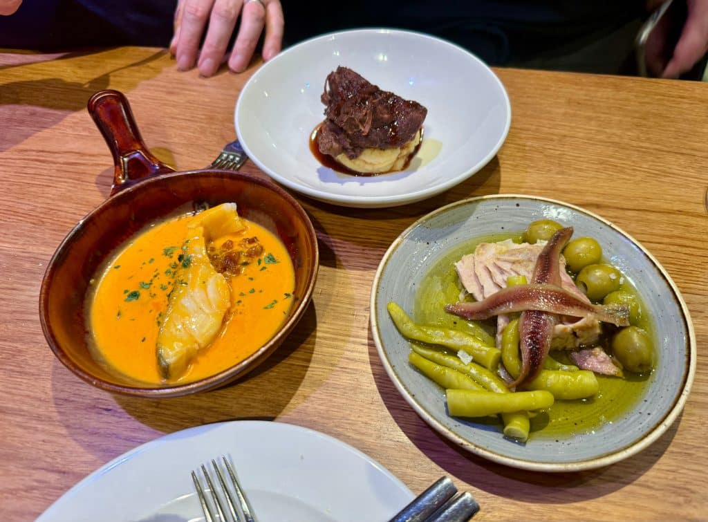 Three tapas plates: a fish dish in orange colored sauce, a piece of meat on mashed potatoes, and a plate of anchovies, hot peppers, and olives.