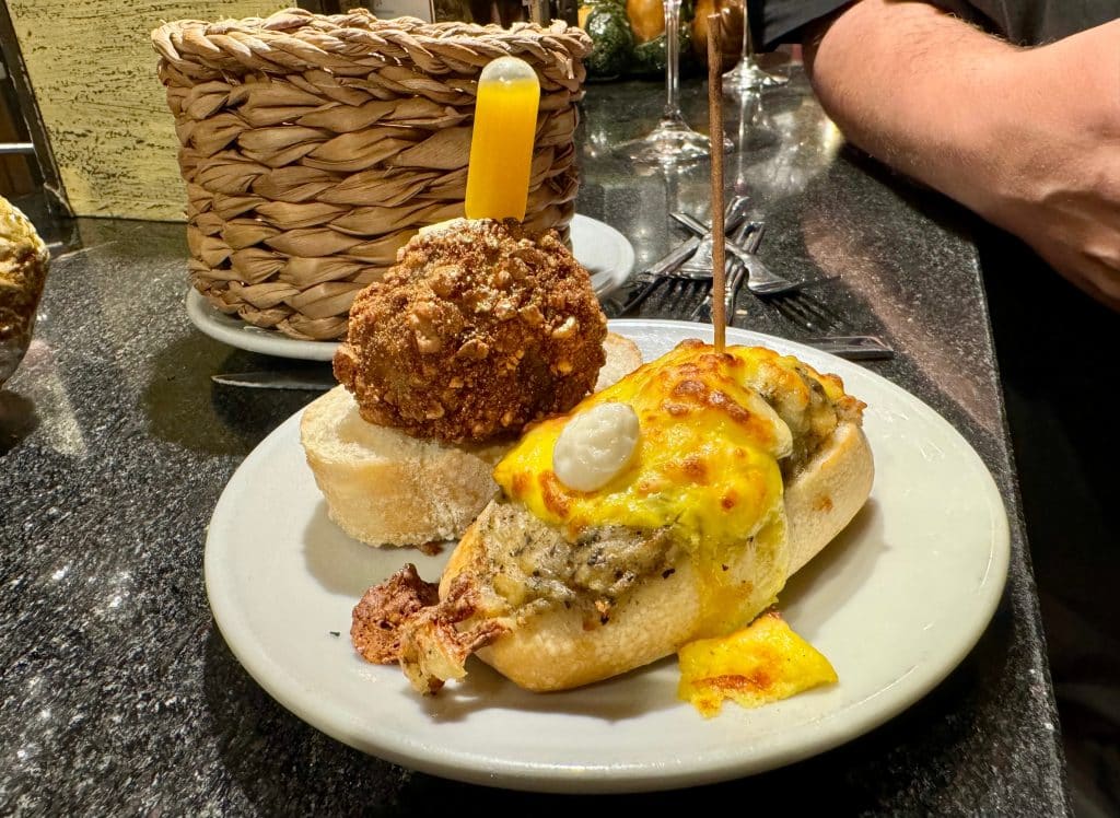 A plate at a pintox bar with a piece of bread and what looks like tuna and melted cheese, next to a piece of bread with some kind of meatball and a plastic tube of a yellow sauce stuck through the top.