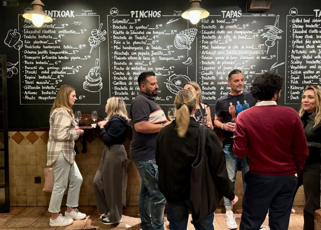 A group of people standing in front of a blackboard menu in a pintxos bar.