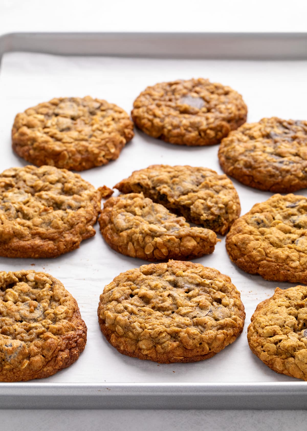 Nine oatmeal cookies on a parchment-lined metal baking sheet, with one cookie broken in half near the center.