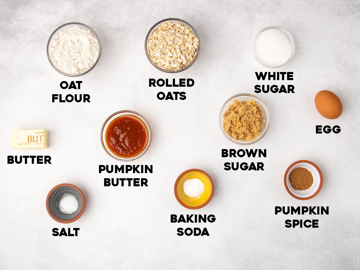 Labeled ingredients arranged on a light gray surface, including oat flour, rolled oats, white sugar, brown sugar, butter, pumpkin butter, salt, baking soda, and pumpkin spice.