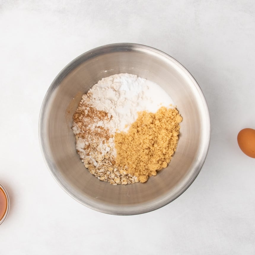 A metal mixing bowl containing unmixed rolled oats, brown sugar, white sugar, and baking soda and light brown powder on a light gray surface, with an egg and a small bowl partially visible nearby.