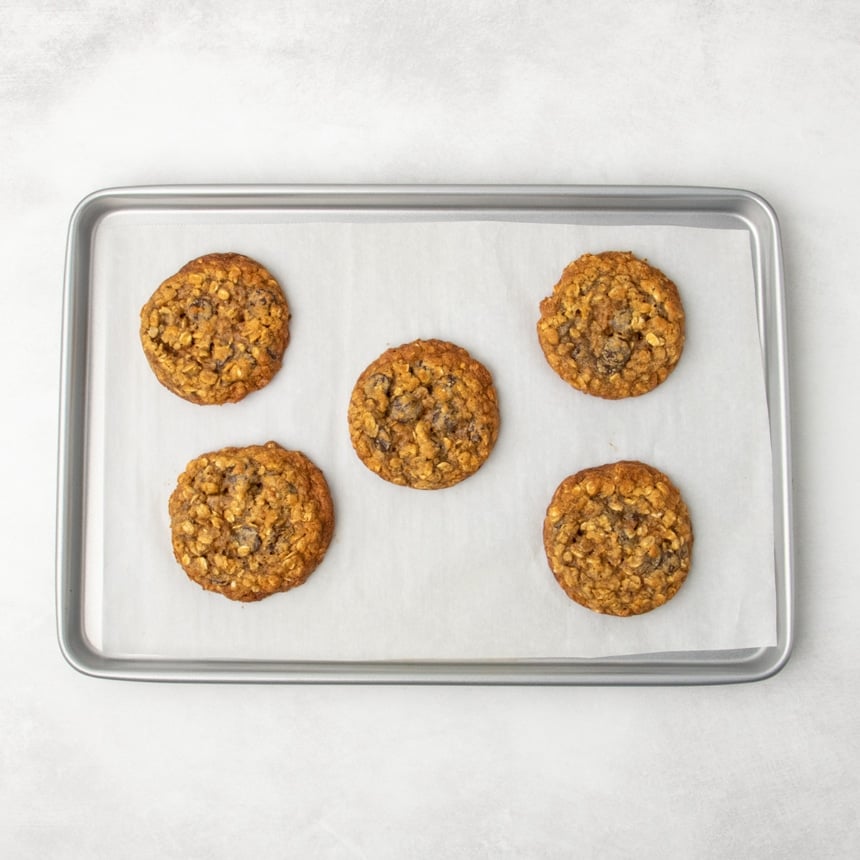 The same parchment-lined metal baking sheet, now with five rust-colored baked pumpkin oatmeal cookies arranged evenly across the surface.