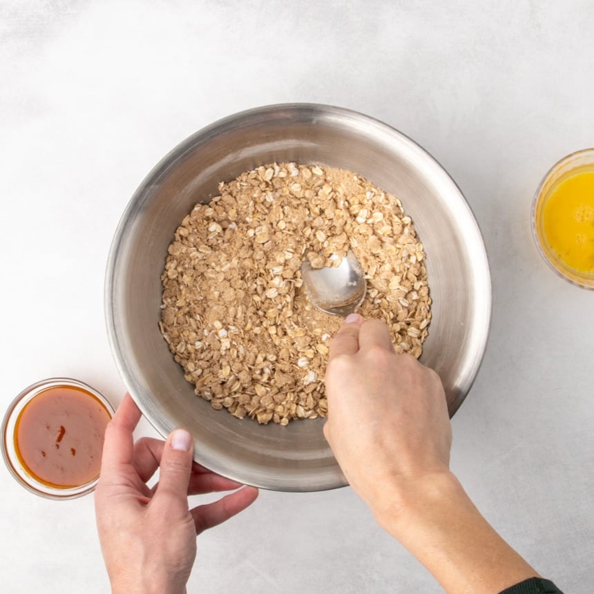 A person’s hands holding a metal spoon while mixing dry ingredients with oats in a metal bowl; small bowls of orange puree and a yellow beaten egg are nearby.