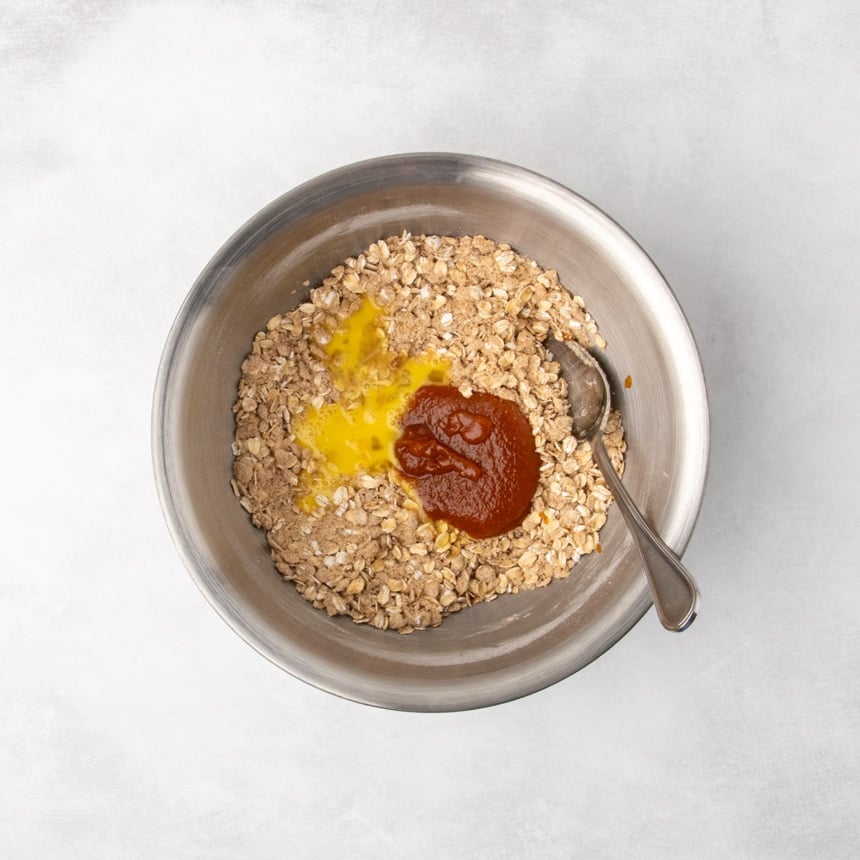 A metal mixing bowl containing a mixture of oats and dry ingredients with a beaten egg and orange pumpkin butter puree added on top, unmixed.