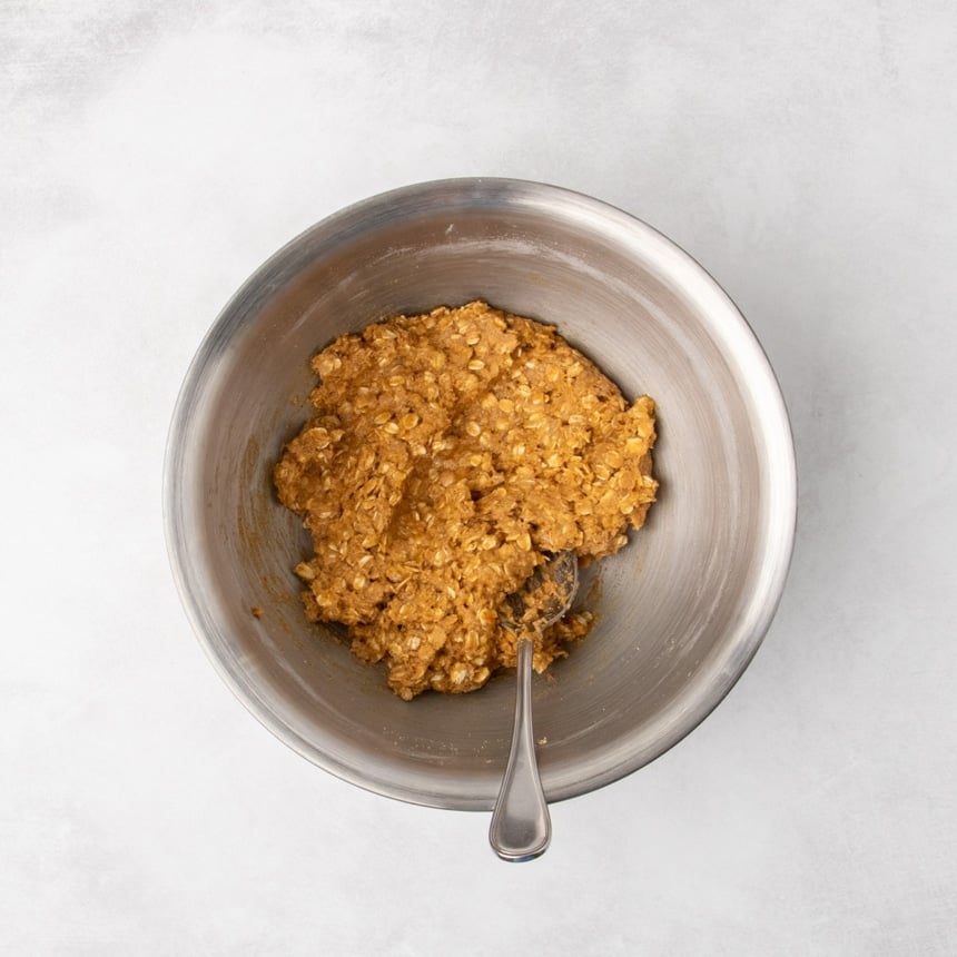 A metal mixing bowl containing combined rusty brown colored raw oatmeal cookie dough with a spoon resting inside.