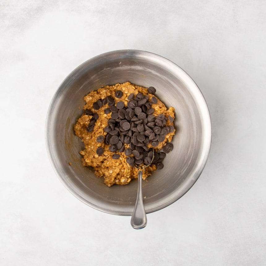 The same metal mixing bowl with the same cookie dough and dark chocolate chips added on top, with a spoon resting in the bowl.