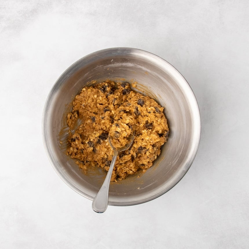 The metal mixing bowl filled with rust colored raw oatmeal cookie dough now mixed with chocolate chips, with a spoon in the bowl.