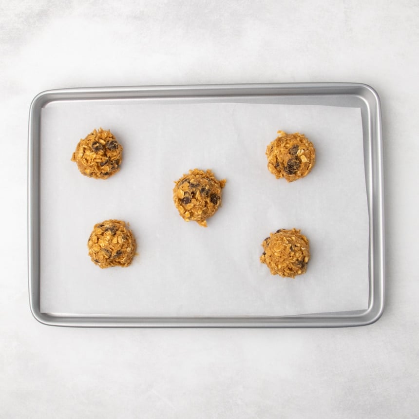 A parchment-lined metal baking sheet with five portions of oatmeal cookie dough spaced evenly apart.